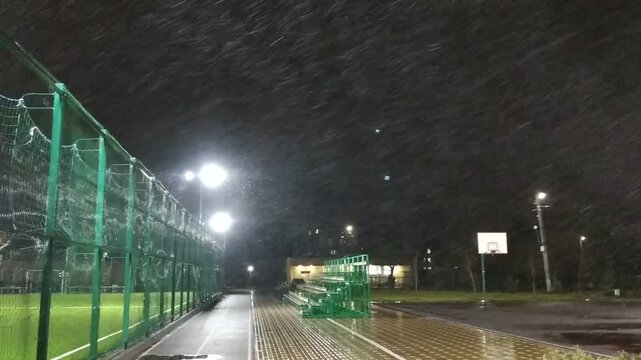 A sports field at night during a heavy snowfall. A soccer field with bright lighting. Green bleachers are visible in the center. The falling snow is clearly visible in the light of the streetlamps.