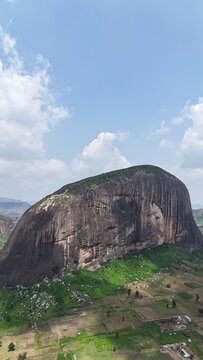 Vertical aerial view of a massive granite rock formation surrounded by green farmland and a sprawling Nigerian landscape