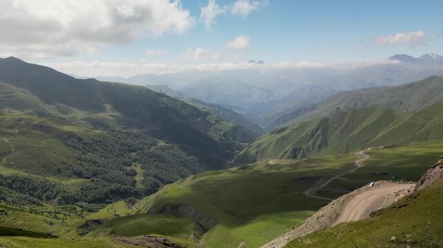 Aerial drone footage looking from Datvisjvari Pass across a deep mountain valley surrounded by green slopes and rugged Caucasus mountains, with clouds covering the distant peaks