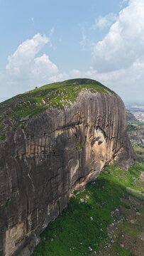 Vertical aerial view of a massive granite rock formation surrounded by green farmland and a sprawling Nigerian landscape