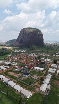 Vertical aerial view of a massive granite rock formation surrounded by green farmland and a sprawling Nigerian landscape