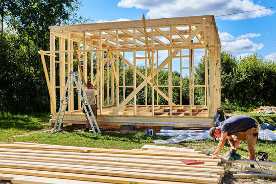 Construction crew of two workers builds timber frame as one cuts boards with circular saw and another fastens braces to wall studs.