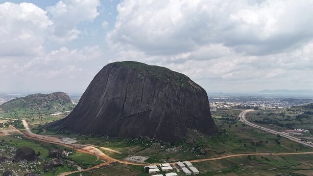 Aerial view of a massive granite rock formation surrounded by green farmland and a sprawling Nigerian landscape.