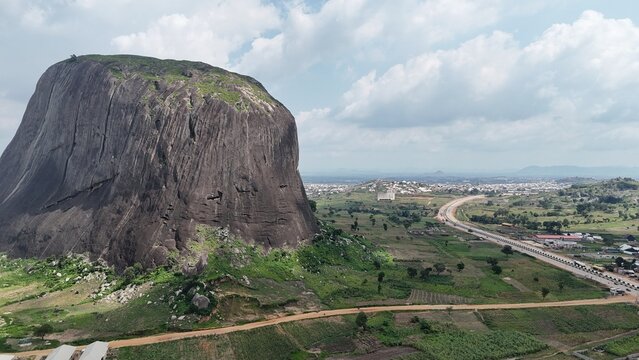 Aerial view of a massive granite rock formation surrounded by green farmland and a sprawling Nigerian landscape.