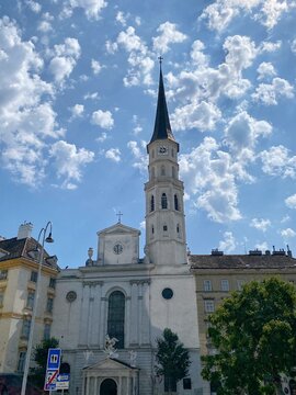 Veduta esterna della storica Chiesa di San Michele (Michaelerkirche) a Vienna con l'alto campanile aguzzo e la facciata neoclassica sotto un cielo azzurro coperto da nuvole bianche