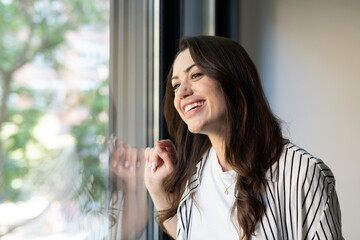 Obraz premium Young woman with a radiant smile standing by a window, looking out with hopeful, relaxed optimism and peaceful contemplation, enjoying natural light and a bright mood