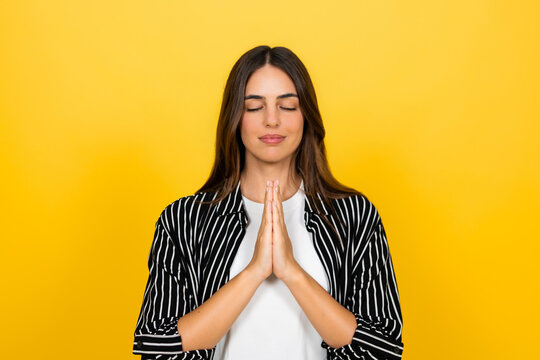 Young woman finding inner peace and practicing mindfulness, closing her eyes and holding hands in a prayer gesture against a vibrant yellow background
