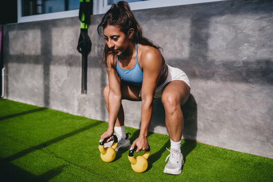 Functional fitness strength training exercise shows Hispanic female athlete squatting to lift yellow kettlebells on artificial grass inside gym room, practicing active wellness goals daily