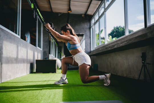 Functional fitness TRX suspension training routine shows athletic Hispanic female athlete holding suspension straps while squatting inside gym, practicing active wellness personal goals daily