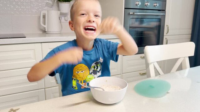 A joyful young boy raises his arms in excitement while sitting at a kitchen table with his bowl of food. He smiles brightly, filled with happiness and playful energy. The modern kitchen is warm