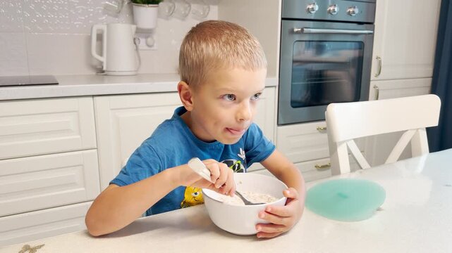 A young boy eats breakfast carefully from a bowl with a spoon. He feels calm and focused as he enjoys his meal. The bright kitchen creates a warm and cozy home setting.