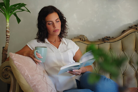 Woman relaxing at home, enjoying leisure time on a luxurious sofa, reading a book, and drinking coffee, illustrating a cozy, peaceful, and comfortable lifestyle