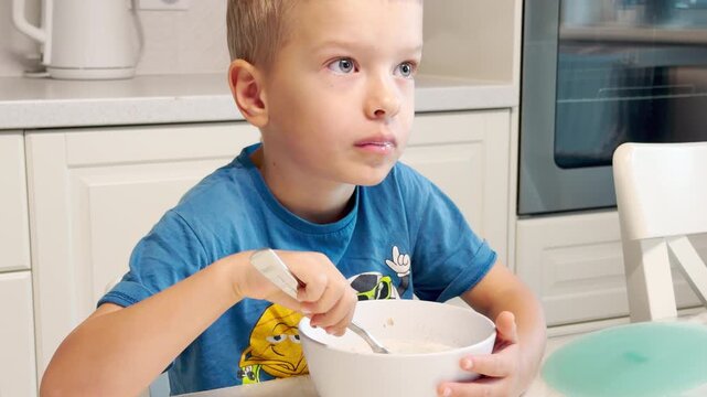 A young boy eats breakfast with a spoon from a bowl. He looks curious and happy in a bright kitchen. His cheerful expression shows morning joy.