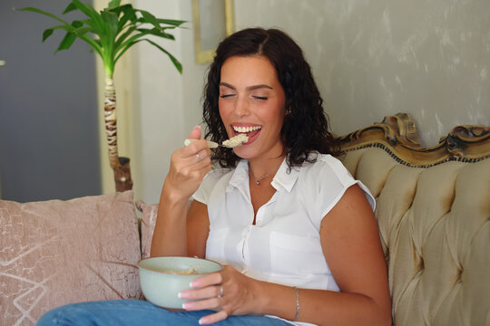 Happy young woman enjoying a healthy breakfast at home, sitting on a sofa and eating a nutritious bowl of oatmeal, embracing a wellness lifestyle with a joyful smile