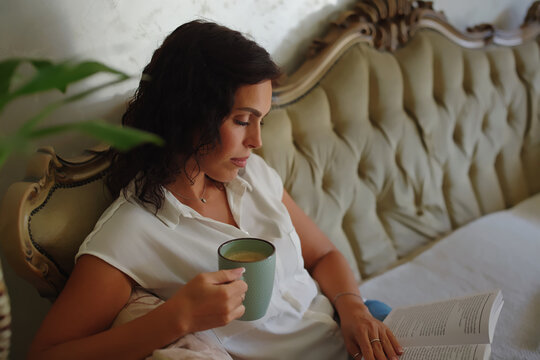 Woman relaxing in a luxurious bed, reading a book and holding a warm mug of coffee, enjoying a cozy, peaceful morning of self-care and quiet leisure at home