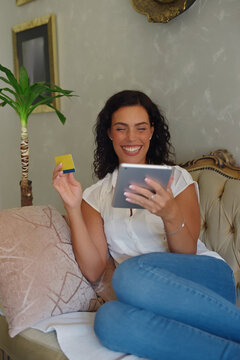 Happy woman relaxing on a couch at home, enjoying online shopping and making secure purchases with a credit card on her digital tablet, smiling and feeling satisfied