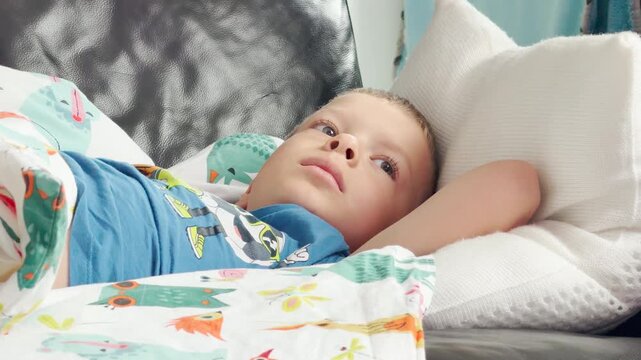 A young boy lies comfortably on a sofa while resting under a colorful patterned blanket. He gazes thoughtfully into the distance with a calm and peaceful expression. The quiet room feels warm and cozy