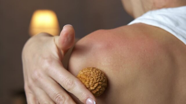 Woman massaging her shoulder with wooden massage ball. Unrecognizable woman massaging her shoulder and back with a spiky wooden ball, performing a self-massage technique for muscle relaxation