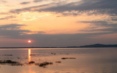 Fototapeta premium Sunrise over a calm lake with reeds and distant hills under a dramatic cloudy sky