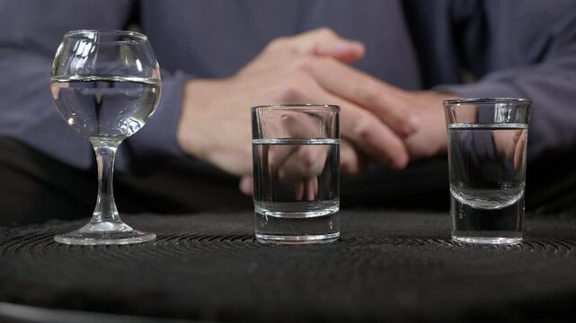 Person hesitating before choosing alcoholic beverage. Nervous man sitting at a dark table with three different glasses of strong alcohol, showing indecision to drinking problems and addiction