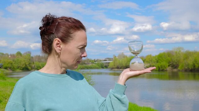 Woman holding an hourglass watching time pass. Serious adult woman holding a sandglass in her palm and watching the sand pour, symbolizing the passage of time and the finite nature of life