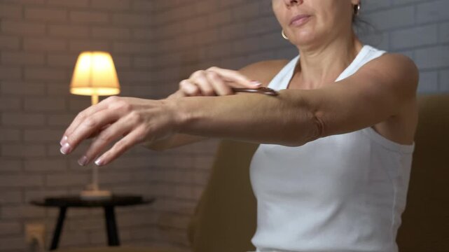 Woman practicing self gua sha arm massage technique. Middle-aged woman in a white tank top performing a gua sha massage on her arm with a scraper, focusing on lymphatic drainage