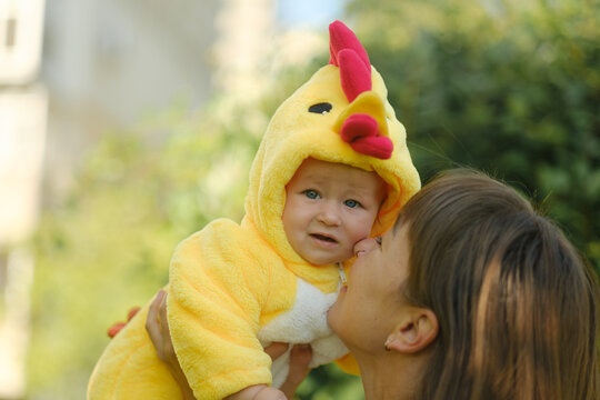Happy mother holding cute baby in yellow chicken costume outdoors in park on sunny day