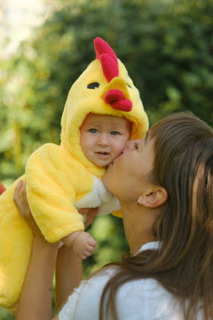 Happy mother holding cute baby in yellow chicken costume outdoors in park on sunny day