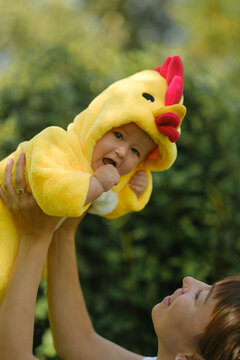 Happy mother holding cute baby in yellow chicken costume outdoors in park on sunny day