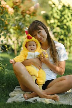 Mother with cute baby in yellow chicken costume sitting on blanket in green park lawn