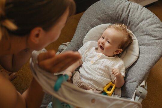 Crying baby boy sitting in bouncer seat at home, upset toddler looking at mother, authentic lifestyle