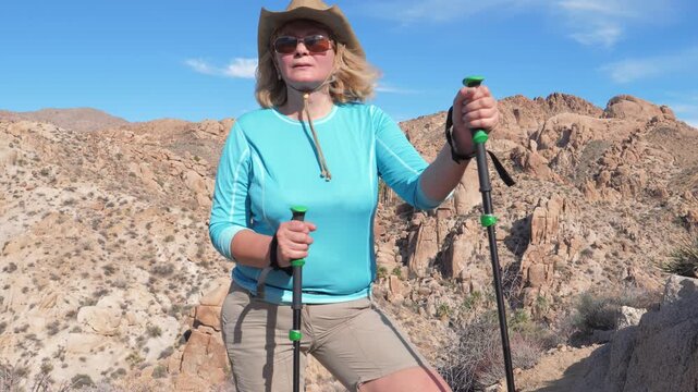 Caucasian woman with trekking poles climbing up steep trail on sunny day, surrounded stunning arid mountain landscape. Tired tourist resting, catching breath and normalizing her breathing after climb.