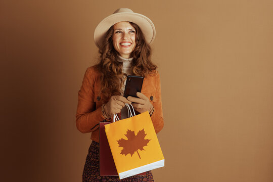 An elegant woman with wavy brown hair and a wide-brimmed hat smiles, holding her smartphone and colorful shopping bags featuring a distinct maple leaf design.