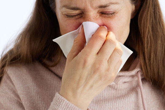 A middle-aged woman in a pink knit hoodie closes her eyes in discomfort while blowing her nose with a white tissue, showing symptoms of a cold or allergy.
