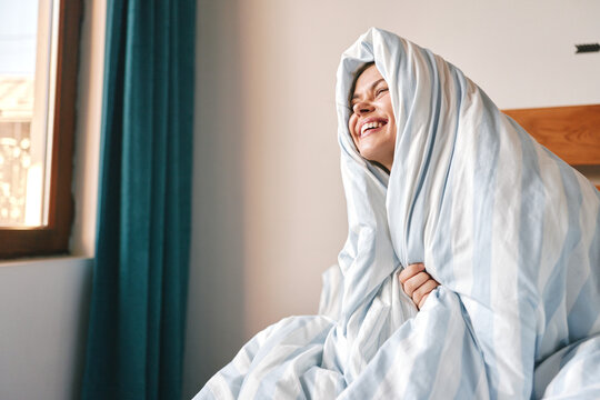 Happy young woman wrapped in a cozy blanket laughs in bed by the window at home, morning mood, comfort and relaxation concept.