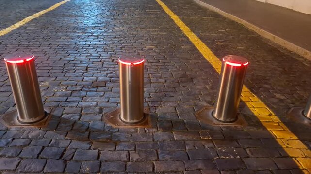 Automatic retractable traffic bollards with red LED illumination glowing on a cobblestone street at night. security barrier, traffic control, pedestrian zone, urban safety.