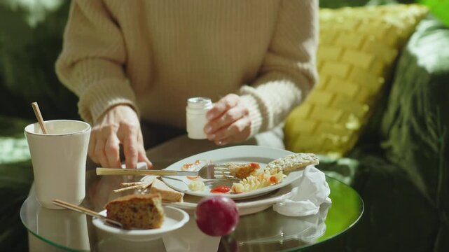 A woman on a green sofa takes a pill from a medicine bottle to find relief from stomach pain and bloating after consuming an unhealthy meal.