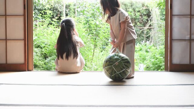 日本の古民家の縁側で、大きなスイカを一緒に運んで笑顔で見つめ合う2人の女の子