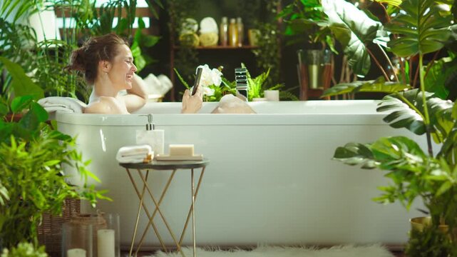 A woman in a bun, wrapped in a towel, smiles looking at her smartphone in a white bathtub, surrounded by lush green plants. This scene captures modern self-care & relaxation in a tranquil home spa.
