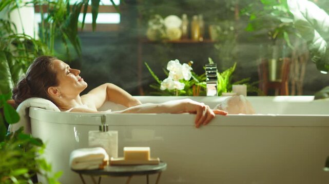 A serene woman with eyes closed & peaceful expression relaxes in a luxurious bathtub, surrounded by lush green plants & steam. This scene creates a tranquil home sanctuary for well-being & self-care.