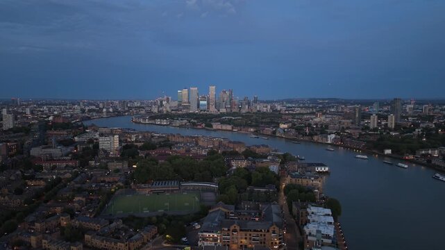 Evening aerial view across London with the River Thames winding through residential districts toward the illuminated skyscrapers of Canary Wharf at dusk.