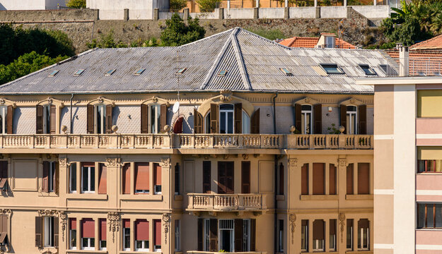 Elegante edificio cl&aacute;sico con amplia terraza, balaustradas, techo a dos aguas con tragaluces y postigos marrones en un entorno urbano.