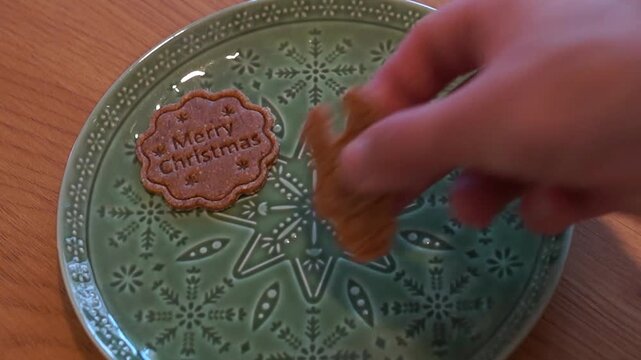 Close-up shot of a man's hand carefully arranging festive Christmas gingerbread cookies (Merry Christmas, reindeer, and gingerbread man) onto a patterned green plate on a wooden table.