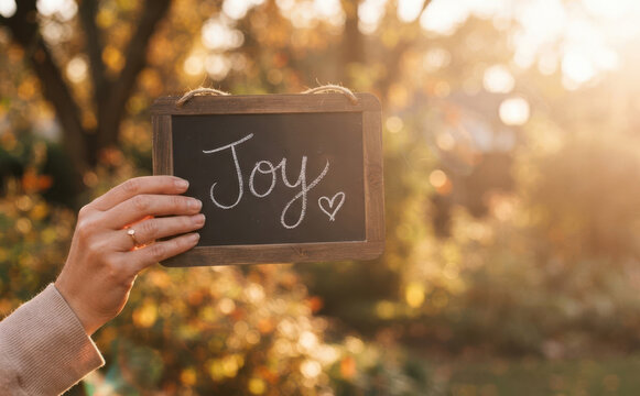 Woman holding a small chalkboard with the word Joy written in chalk outdoors in autumn representing happiness, positivity, inspiration, and emotional well-being.