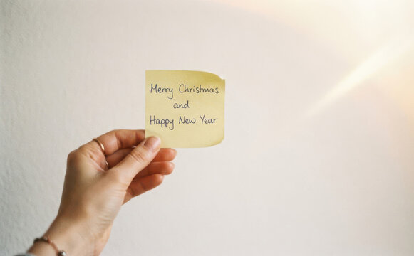 Hand holding a yellow sticky note with Merry Christmas and Happy New Year handwritten message for winter holiday greeting concepts