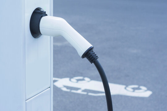 Electric vehicle charging cable handle plugged into power station with ev parking slot sign in background. Green energy transportation, eco friendly technology and sustainable mobility concept.