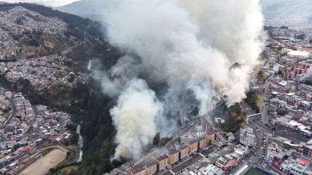 Aerial view of a large fire and thick smoke rising above Quito urban neighborhoods, roads, sports fields and a wooded ravine in Ecuador.