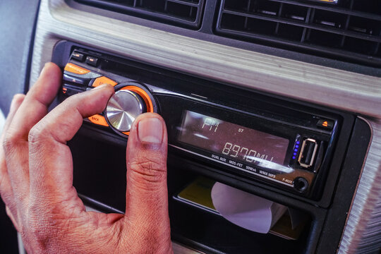 Close-up of a person's hand turning the volume knob of a car head unit stereo player, tuning the FM radio station inside a vehicle dashboard.