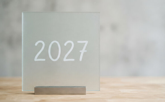 Minimalist frosted glass sign with the year 2027 printed on a wooden table representing future planning and new year goals