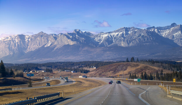 Scenic asphalt highway leading toward snow capped Rocky Mountains with cars driving under a blue sky in winter